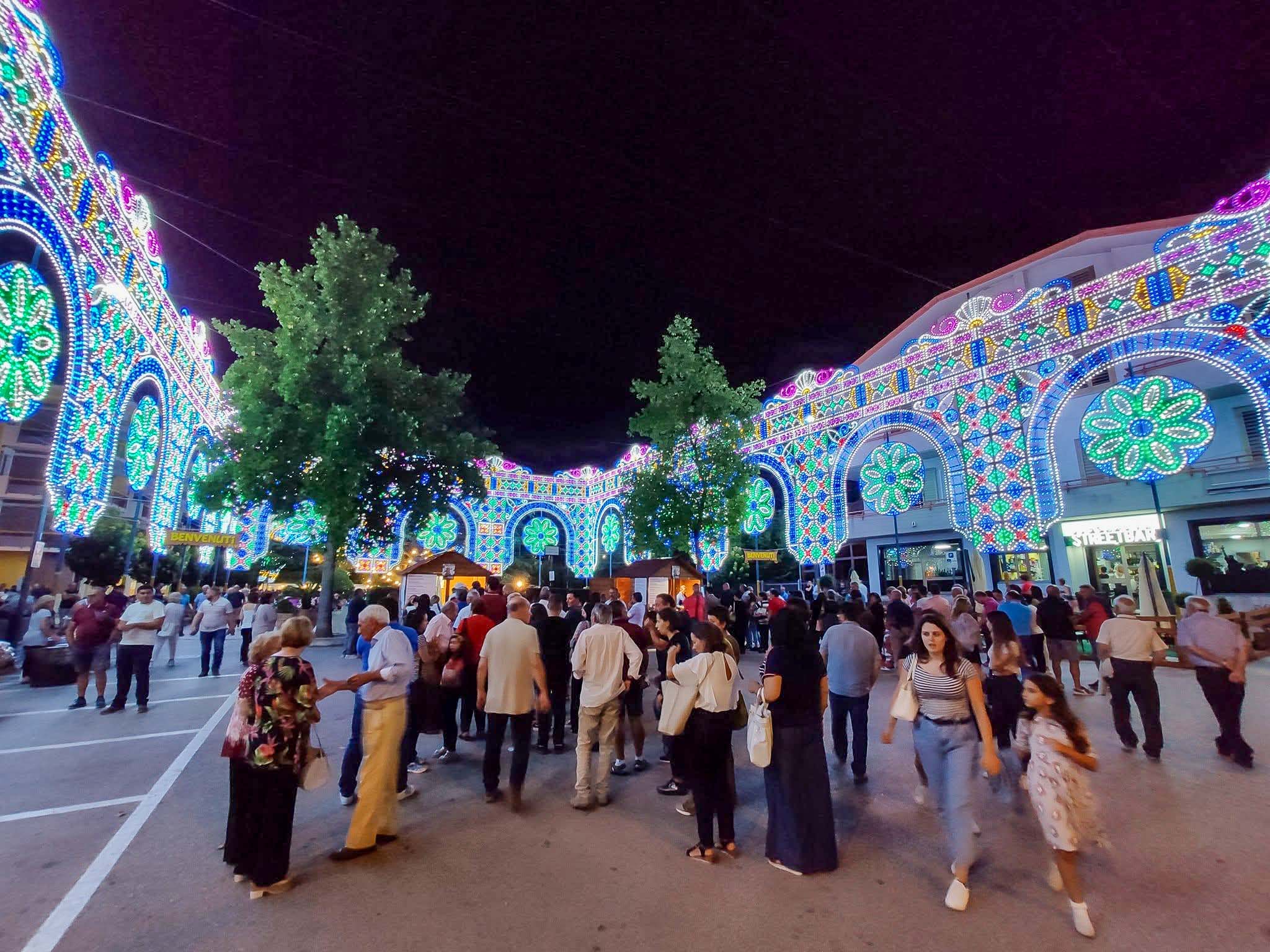 Piazza gremita di persone sotto la maestosa galleria di luminarie ad arco accese a festa